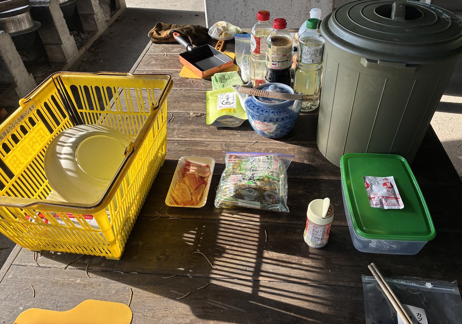 屋外の調理台に並んだ卵焼き器、しょうゆ類、漬物のパック、黄色いカゴ